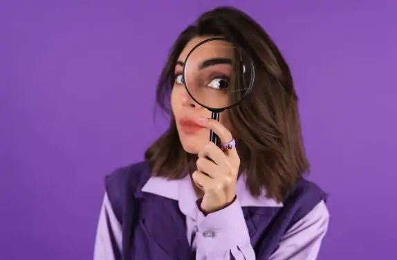 Young woman in shirt and vest on purple background having fun with magnifying glass in hand